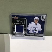Jake Virtanen Rookie Materials Jersey hockey card front showing the player's image, Vancouver Canucks uniform, and embedded navy blue jersey piece.