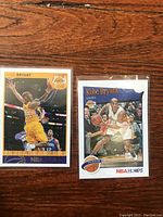Front view of two Kobe Bryant NBA Hoops basketball cards in protective sleeves, one showing him in a yellow Lakers jersey and the other in white.