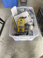 Plastic bin containing various electronic accessories, showing Sony battery pack prominently
