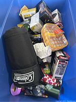 Various items cluttered in a blue bin including Coleman fabric item, orange and white plastic container, numerous boxes and tumblers.