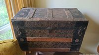 Front and side angled view of antique steamer trunk showing embossed floral tin panels, black wood frame, metal lock, latches and leather handle.