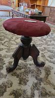 View of antique adjustable wood stool with carved legs and burgundy padded top taken from a side angle on a patterned carpet.