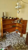 Wide angle image showing the corner bookcase with three wood shelves and a vertical side shelving with curved edges, placed against a wall with various items on shelves (not included).