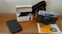 Photo of Honeywell Electric Eye 35 camera with brown case, Braun Hobby F30B flash unit in its box, and a black pocket calculator on wooden surface.