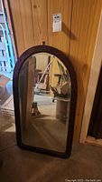 Full view of the arched wood framed mirror leaning against a wooden paneled wall in a basement with visible mold on the frame.