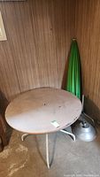 Full view of round patio table with paint fading and umbrella closed standing nearby with metal base on floor.