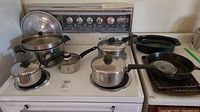 Multiple cooking pots, frying pans, baking pan, and small roaster tray arranged on a stove and nearby surface, showing overall lot contents.