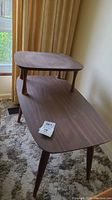 Vintage two-tiered side table with a larger bottom tier and smaller top tier, made of laminate with visible wood grain and scratches, standing on tapered wooden legs
