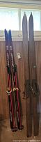 Both pairs of skis, wooden and Jarviner, side by side angled against a wall under a window.