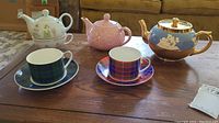 Set of four teapots and two cup and saucer sets displayed on wooden table.