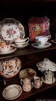 Six assorted teacups with saucers, various floral and gold patterns, alongside decorative cake plates and dessert plates on a dark wooden shelf