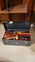 Metal toolbox open on floor showing assortment of tools inside, including mallets, hammer, tape measure, pliers, and screwdrivers.