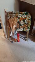 Two metal TV tables with floral fabric tops, one beige umbrella, one wooden cane with a white handle, and a small red container of matches grouped together on carpeted floor.