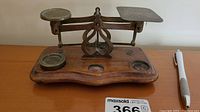 Front view of the vintage postal scales showing wooden base, brass balance arms, one circular weight on the left, and the square inscribed scale plate on the right.