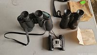 Three pairs of binoculars shown on a table: one vintage pair with tan carrying case, two modern black binoculars including a compact model.
