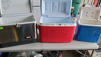 Photo of three coolers on table: large metal Coleman cooler, medium red Coleman plastic cooler, smaller blue Igloo plastic cooler