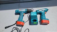 Photo showing two cordless drills and a charger laid flat on a surface, both drills with orange battery packs, charger in the middle.