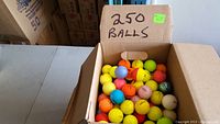 Box labeled with '250 BALLS' filled with assorted colored golf balls including yellow, orange, blue, pink, green, and white.