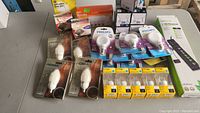 Table with assorted packaged light bulbs including candle shaped LED bulbs from Nova, standard Philips bulbs, multiple Luminus LED bulbs in clear boxes, all new unopened. Also a boxed power bar.
