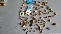 Overhead photo showing many assorted pins spread on a surface. Includes pins in various shapes and designs, several duplicates visible.