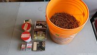 Wide view showing orange pail with spiral nails and several small boxes and containers on table.