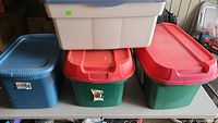 Four plastic storage totes on a table, including one large blue Sterilite tote, one white tote, and two green totes with red lids.