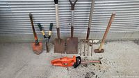Full view of all yard tools arranged on the ground showing the hedge trimmer, shovels, fork, sledgehammer, grass clippers, and spades against a corrugated metal background.