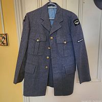 Front view of WW2 RCAF blue wool coat hanging showing multiple pockets, brass buttons and patches on shoulders and sleeve.