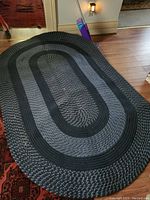 Full view of an oval braided jute and fabric area rug placed on a wooden floor near a doorway with visible wear and dirt.