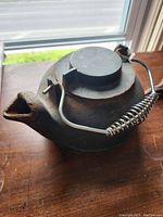 Side view of cast iron kettle showing spout, round body, lid, and coiled wire handle resting on a wooden surface near a window with daylight.
