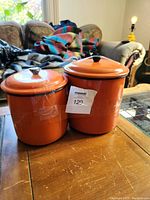 Two orange enamel cylindrical kitchen storage canisters with lids on wooden surface, in living room setting.