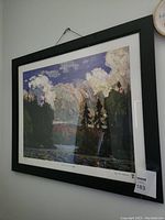 Angled front view of framed print showing image of landscape with trees, water, and sky with clouds.
