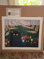 Front view of the lithograph showing three black cats with yellow eyes amidst colorful flowers and trees, mounted on cardboard with label.