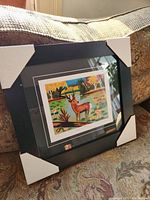 Framed fine art print displayed on a couch with protective corner guards on the frame.