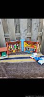 Full view of open vintage Fisher Price fold-out house with multiple rooms and furniture, five Little People figures on the shelf above, and three toy vehicles lined up below the house.