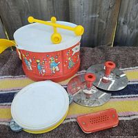 Photo of the vintage Fisher Price toy drum with yellow drumsticks, two silver toy cymbals with red handles, and a red toy harmonica on a striped fabric surface.