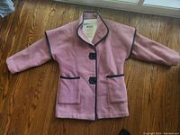 Full front view of pink wool vintage jacket with black trim and two front pockets laid flat on wooden floor under window light.