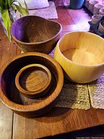 Photo showing five wooden bowls of varying sizes and wood tones, displayed on woven placemats on a wooden floor, with plant and pots in background.