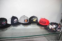 Front view of six baseball caps on glass shelf showing diverse brand logos and designs
