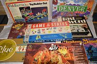 Photo showing the eight boxed vintage board games arranged with titles visible, including Game of the States, Clue, Sly, Mystery Mansion, Last Word, Denver In-A-Box, Gold 'N' Glory, and Fiddlesticks Toy Builder Set.