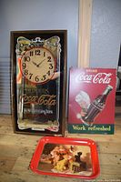 Photo showing the Coca-Cola wall clock, metal sign, and serving tray arranged together on a wooden floor.