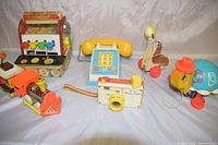 Photo showing all six vintage Fisher Price toys including cash register, telephone, pull seal and turtle, camera, and train with car on light gray fabric background.