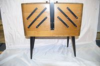 Closed view of a wooden foldout sewing box on angled legs with a black handle and metal hinges.
