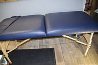 Blue foldable massage table with wooden legs, set up for use on wood floor.