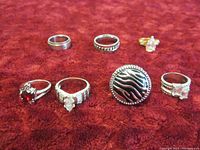 Seven sterling silver rings arranged on a red textured surface showing variety including plain bands, faceted stones, and a zebra patterned ring.