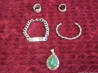 Group photo of sterling silver jewelry assortment on red textured background showing two rings, two bracelets, and a pendant with green stone