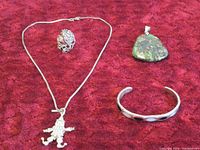 Four pieces of jewelry on a red textured fabric background: a silver chain necklace with clown pendant, silver ring, silver cuff bracelet with dark stones, and pendant with green semi-precious stone.