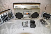 Front view showing all vintage radios, Sony boombox, two handheld portable devices, small TV, and accessory cables.