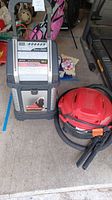 Pair of items; Vector digital battery charger standing next to a red Shop Vac 5-gallon wet/dry vacuum on garage floor.