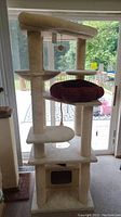 Full view of tall beige cat tree showing multiple platforms with maroon cushion on middle platform and hanging toy ball under top perch